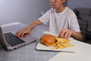 How to get kids to eat healthy foods. Image of 12 year old boy eating burger in fries in front of laptop.